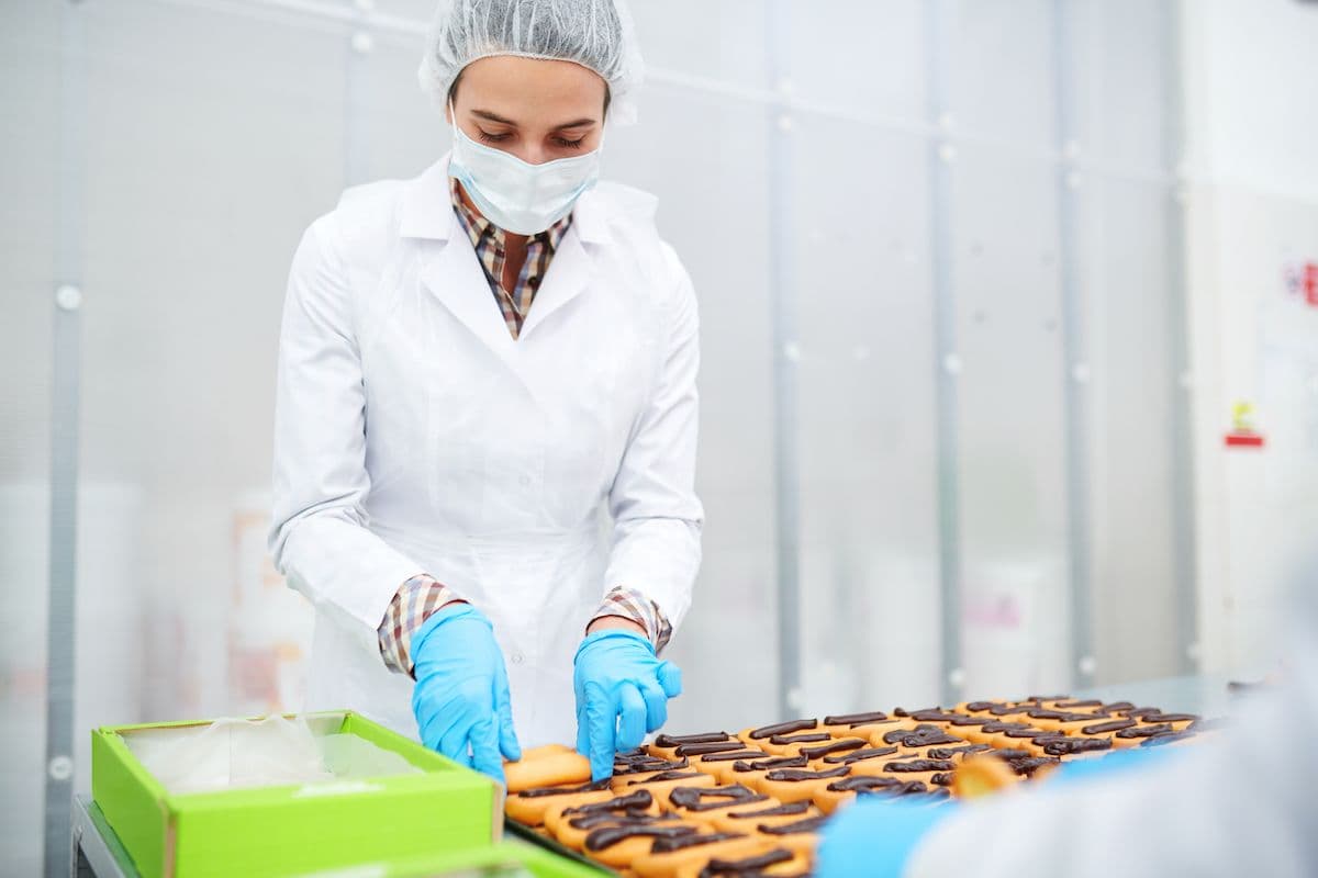 Food handler in a hairnet, mask and gloves packing pastries on a production line